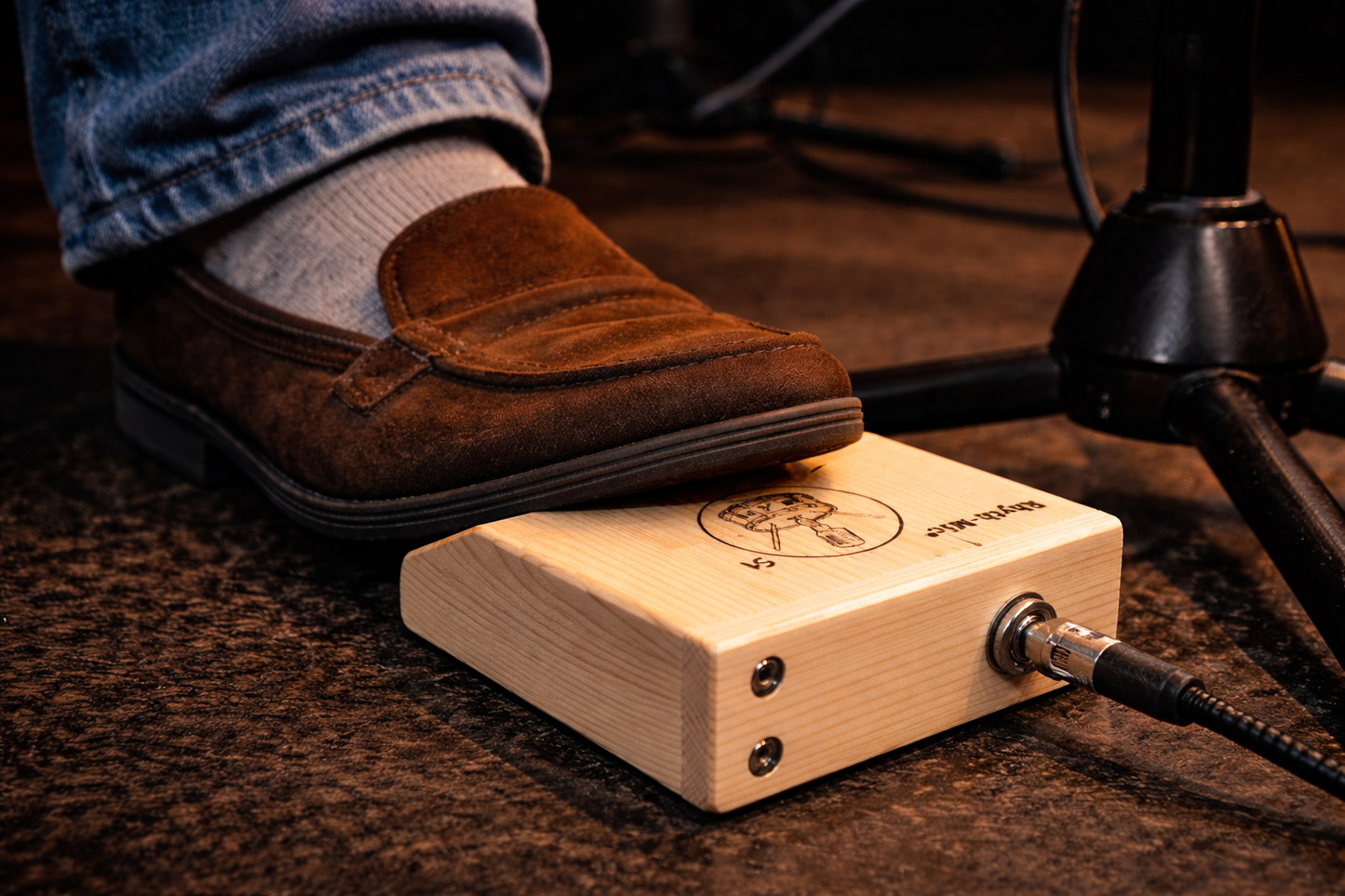 Photo of a rhyth-mic S1 snap stomp box under a musicians foot, plugged into an instrument lead. A microphone stand is visible in the picture.
