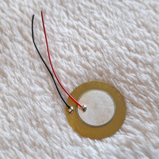 close up of a circular brass piezoelectric pickup with approx 5cm of live and earth wire attached. on a white background