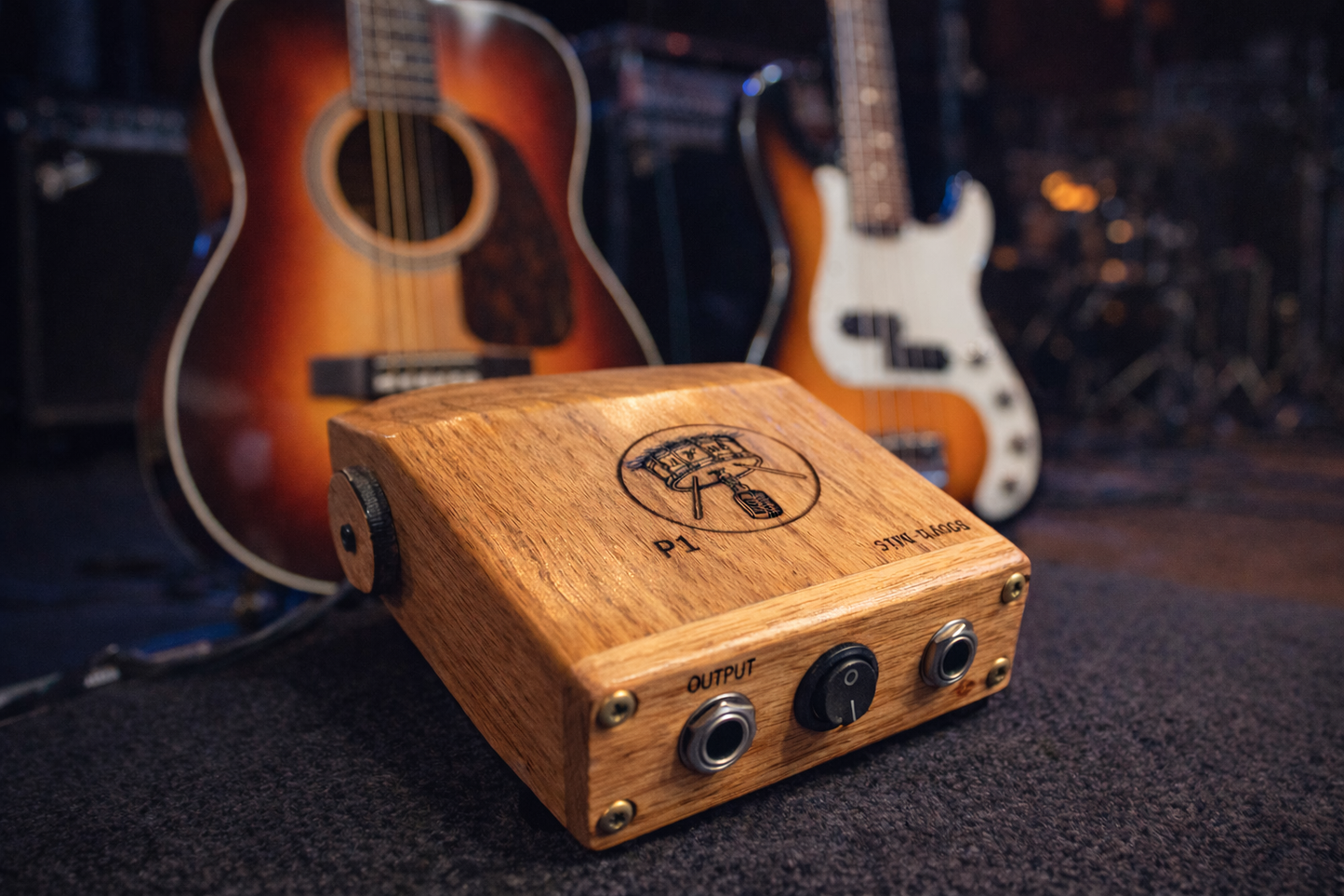 Rhyth-mic P1 play-through stompbox posed attractively on stag in front of an acoustic guitar and an electric bass.